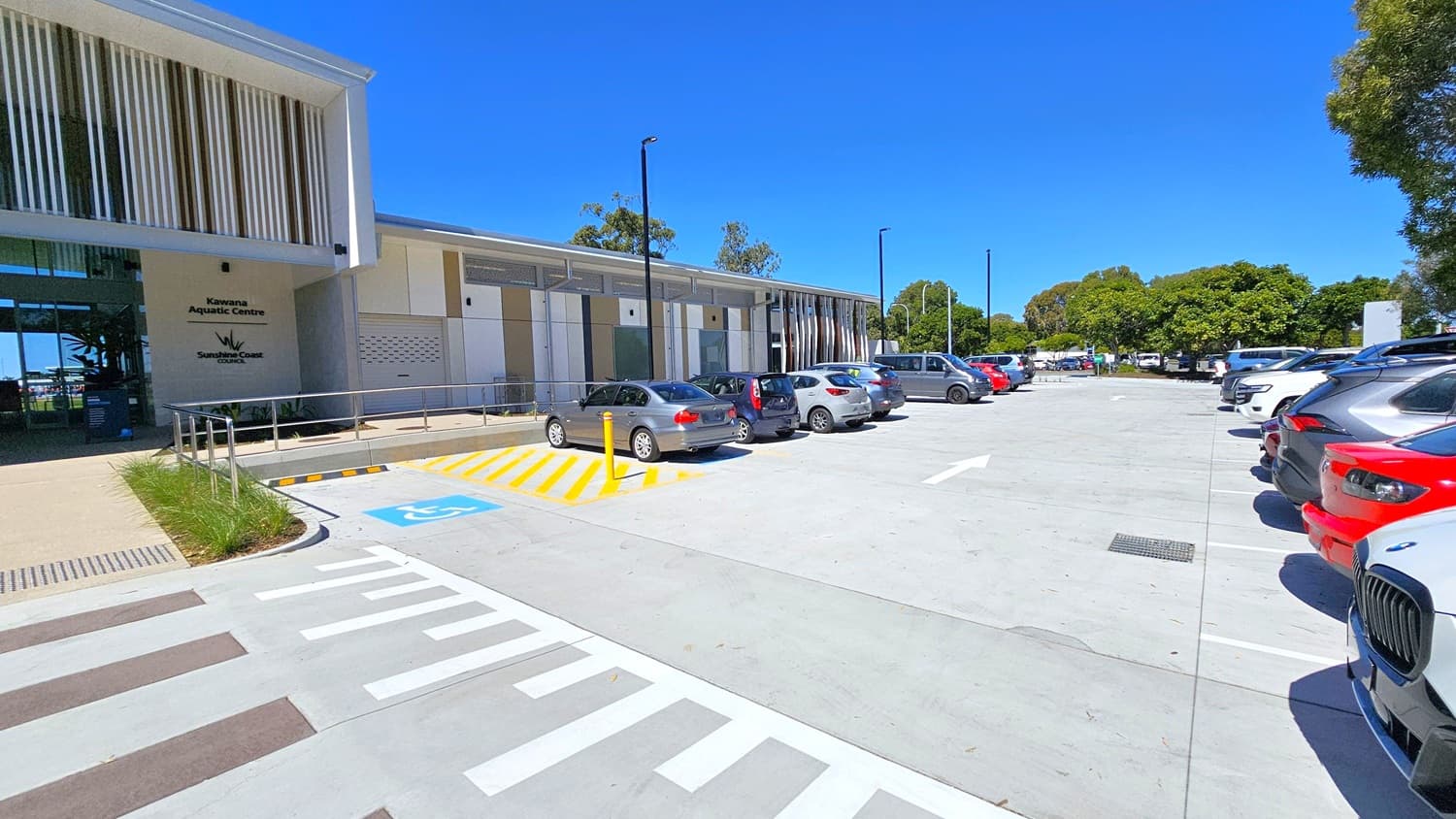 On the left, a modern building with vertical slats and signage for Kawana Aquatic Centre. In the middle, a row of parked cars, including a silver and a red car, with a marked disabled parking space in front. On the right, more parked cars and a line of trees in the background. The ground is a light grey concrete with white and yellow markings.
