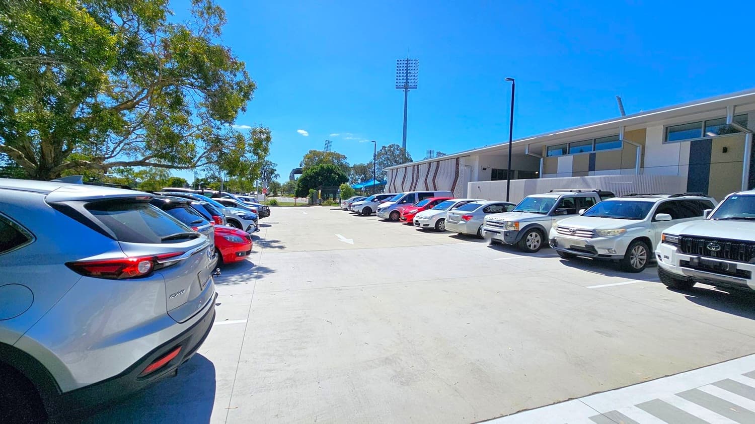 Cars are parked in two rows on either side of a concrete parking area. On the left, a row of vehicles is shaded by a large tree with dense foliage. On the right, another row of cars is parked alongside a modern building with a flat roof and large windows. In the background, tall stadium lights and more trees are visible. The ground is paved, and the sky is clear.