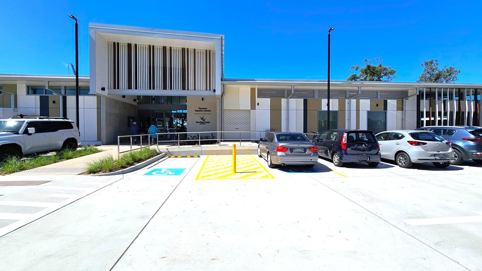 Modern building with a flat roof and vertical slats on the facade. In front, a car park with several vehicles, including a designated disabled parking space marked in blue. A ramp with metal railings leads to the entrance. The ground is paved with concrete, and there are small patches of grass near the walkway. Tall streetlights are positioned along the parking area. Trees are visible in the background.