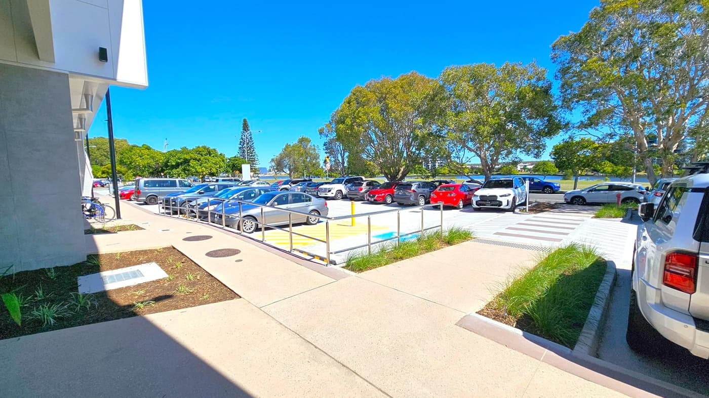 A car park with various vehicles, including silver, red, and white cars, is visible. On the left, a building with a grey wall and a bicycle is parked nearby. A pathway with a railing runs alongside the parking area. Trees with green foliage are in the background, and a tall, narrow tree is visible in the distance. The ground is paved, and there are small patches of grass near the pathway.