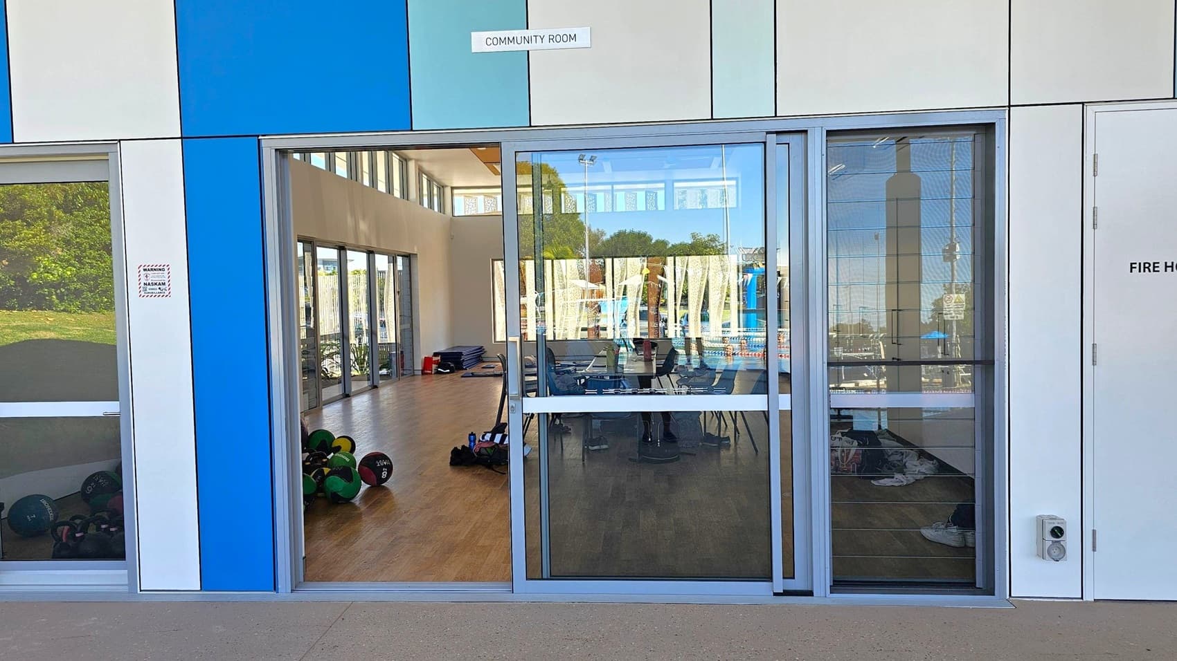 A building exterior with a blue and white panelled facade. In the centre, glass doors lead into a room labelled "Community Room." Inside, exercise equipment like medicine balls is visible on a wooden floor. Large windows line the interior, allowing natural light. To the right, a door marked "Fire Hose" is present. The reflection in the glass shows outdoor elements, including trees and a pool area. The ground outside is concrete.