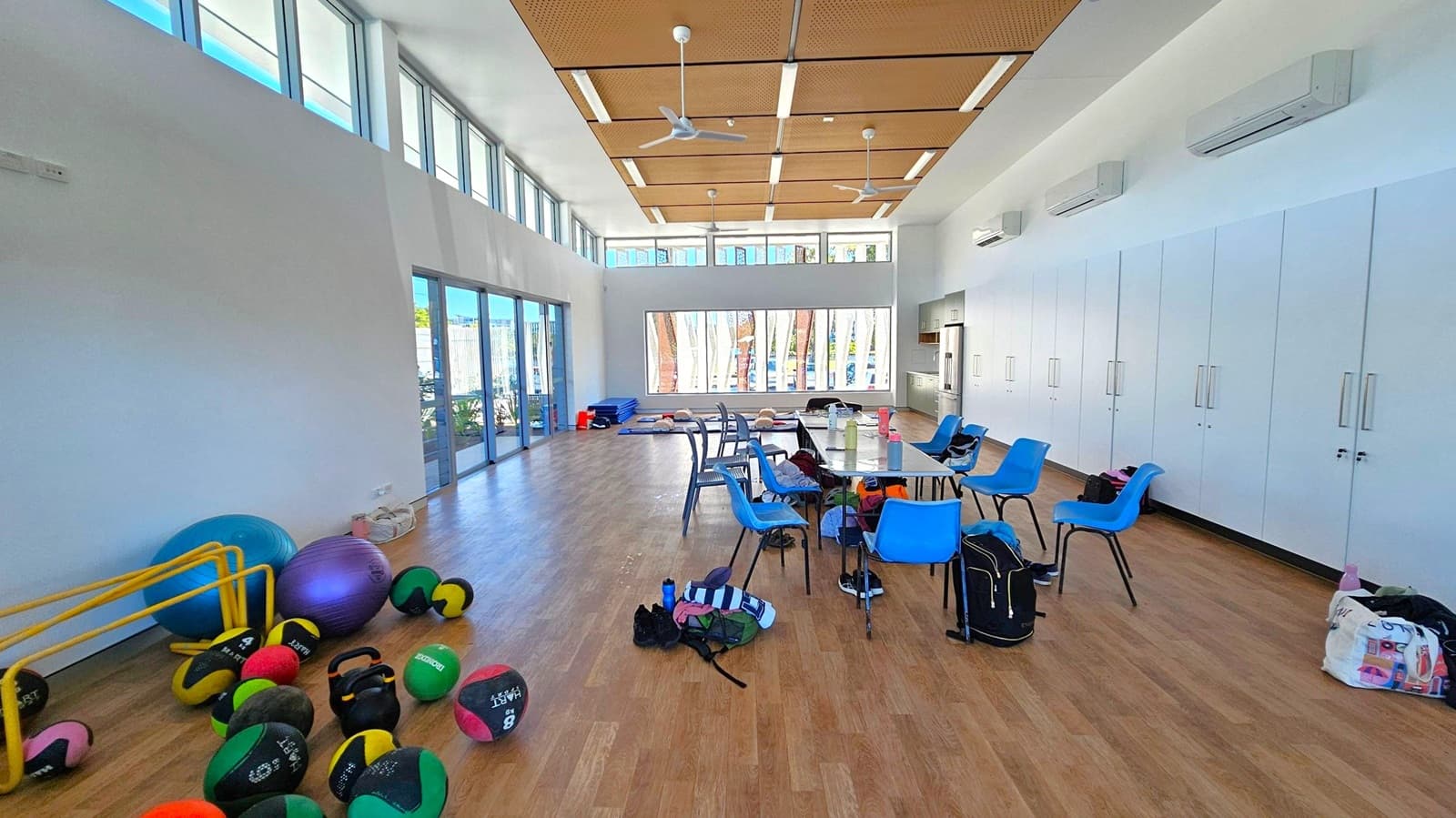 A spacious room with wooden flooring and a high ceiling featuring wooden panels and ceiling fans. On the left, there are large windows and exercise equipment, including medicine balls and a balance ball. In the centre, a table surrounded by blue chairs, with bags and water bottles scattered around. The right side has white cabinets and a kitchenette area. A large window at the back provides natural light, and air conditioning units are mounted on the walls.
