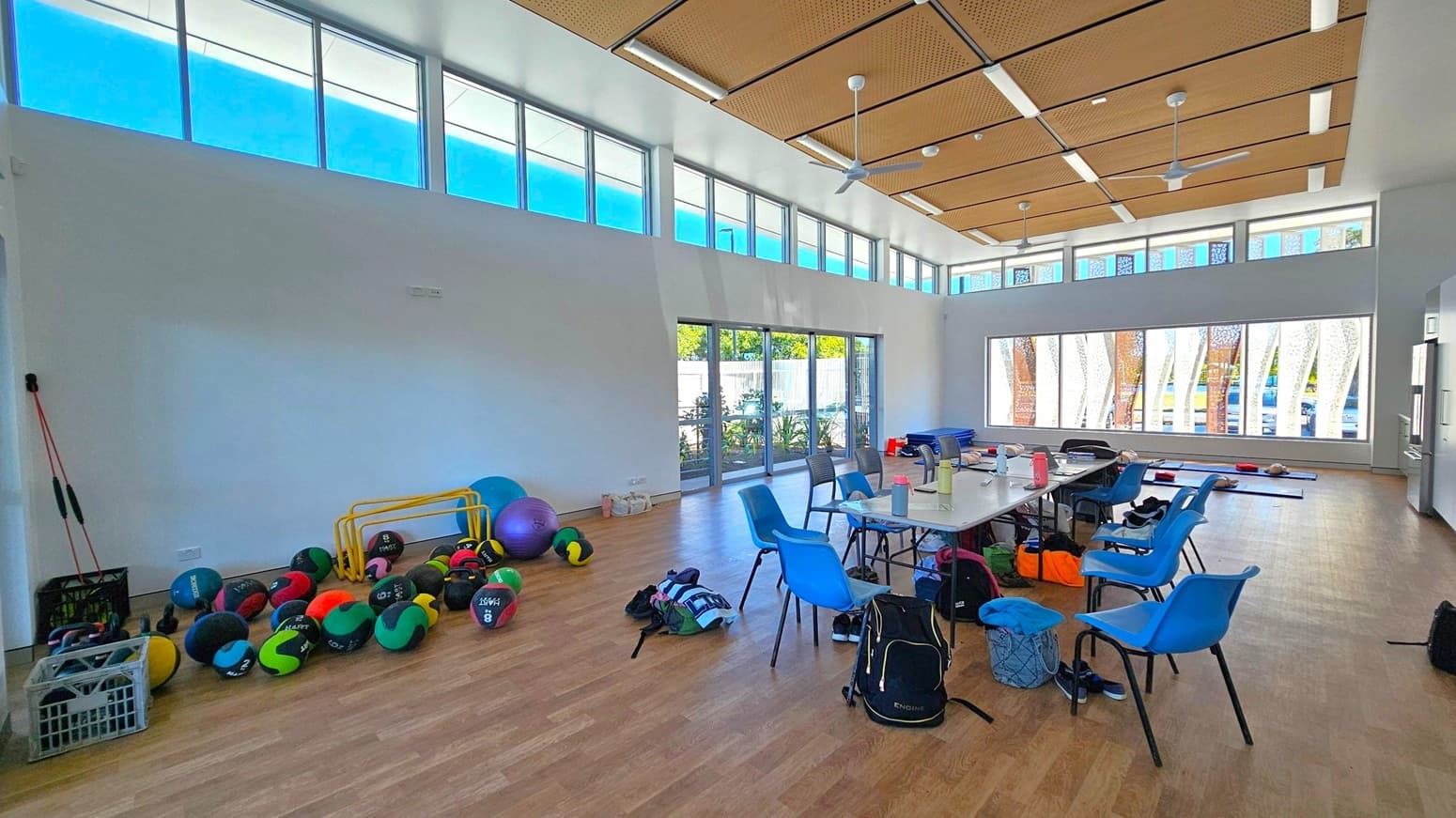 A spacious room with a wooden floor and a high ceiling featuring wooden panels and ceiling fans. On the left, there are various exercise balls and equipment, including colourful medicine balls and yellow hurdles. In the centre, a table is surrounded by blue chairs, with bags and water bottles scattered around. The right side has large windows with patterned designs, allowing natural light to illuminate the space. The background shows greenery outside through the glass doors.