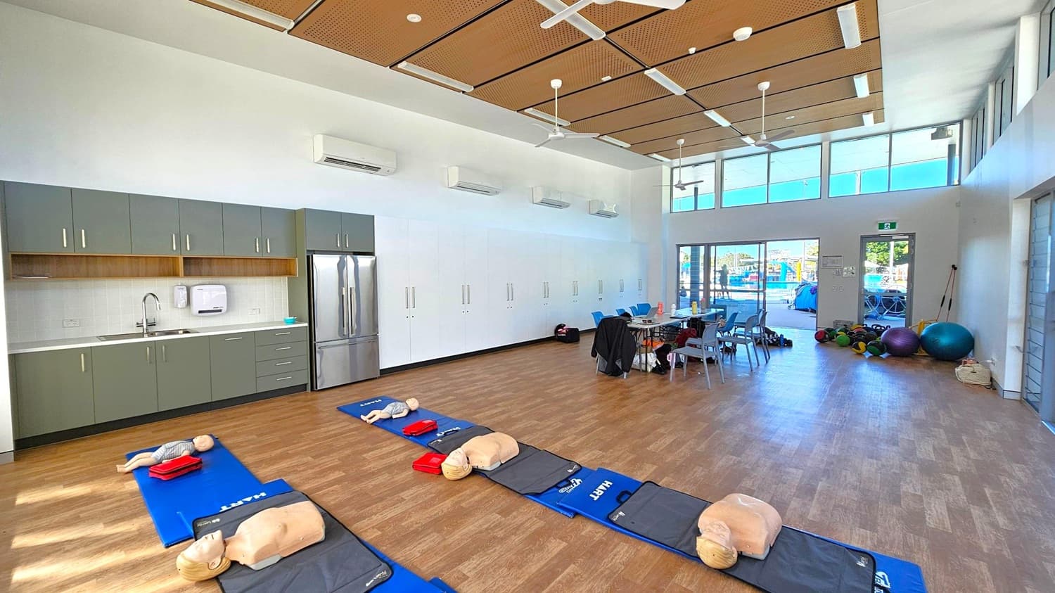 A spacious room with a wooden floor and a high ceiling featuring wooden panels and ceiling fans. On the left, there is a kitchenette with grey cabinets, a sink, and a stainless steel refrigerator. In the centre, CPR mannequins are placed on blue mats. On the right, a table with chairs is surrounded by exercise balls and equipment. Large windows and a glass door provide natural light, with a view of an outdoor area.