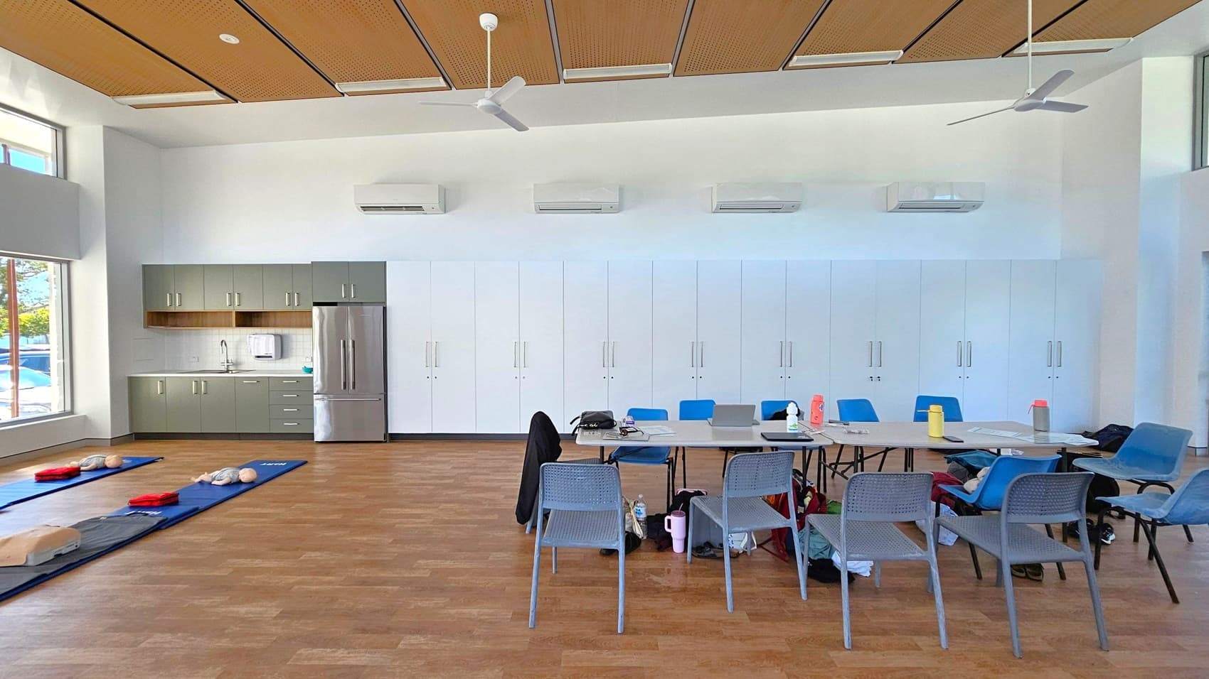 A spacious room with wooden flooring and a ceiling featuring wooden panels and ceiling fans. On the left, there are large windows and a kitchenette with green cabinets, a sink, and a stainless steel fridge. In the centre, a table is surrounded by blue and grey chairs, with various items like water bottles and bags on and around it. On the right, white cabinets line the wall. Blue mats with CPR mannequins are laid out on the floor.