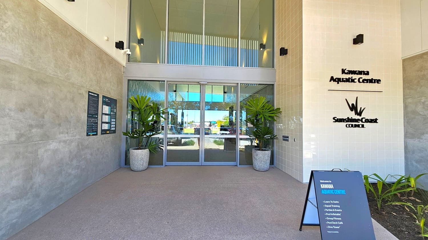 Entrance to a building with glass doors in the centre, flanked by large potted plants. The left wall is grey with informational signs, while the right wall is white with the text "Kawana Aquatic Centre" and "Sunshine Coast Council". A black sandwich board on the right lists activities like "Learn to Swim" and "Squad Training". The floor is grey concrete, and the ceiling is high with recessed lighting. Outside, a glimpse of greenery and a clear day is visible through the glass.