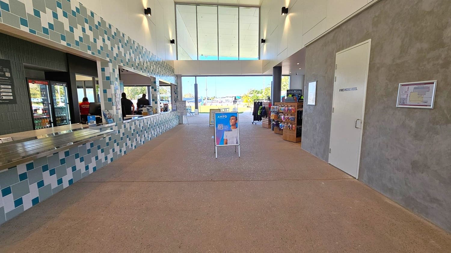 A spacious indoor area with a tiled counter on the left, featuring a mosaic pattern in shades of blue and grey. Behind the counter, there are windows with people visible inside. On the right, a grey and green textured wall with a door labelled "Fire Hose Reel" and a wall-mounted map. In the centre, a sign with a person’s image stands on a speckled floor. The background shows large glass windows and doors leading outside, with a view of trees and a clear sky.