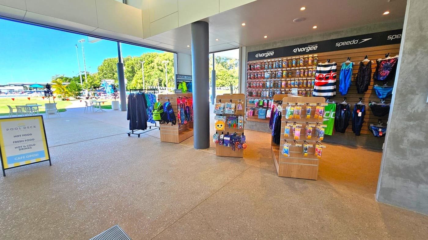 Inside a shop with a polished stone floor, there are several wooden display stands in the centre showcasing swimwear and accessories. On the right, a wall features hanging swim gear and branded items. A large window on the left reveals an outdoor area with tables, chairs, and a view of a swimming pool surrounded by greenery. A sign near the entrance advertises food and drinks. The ceiling is modern with recessed lighting.