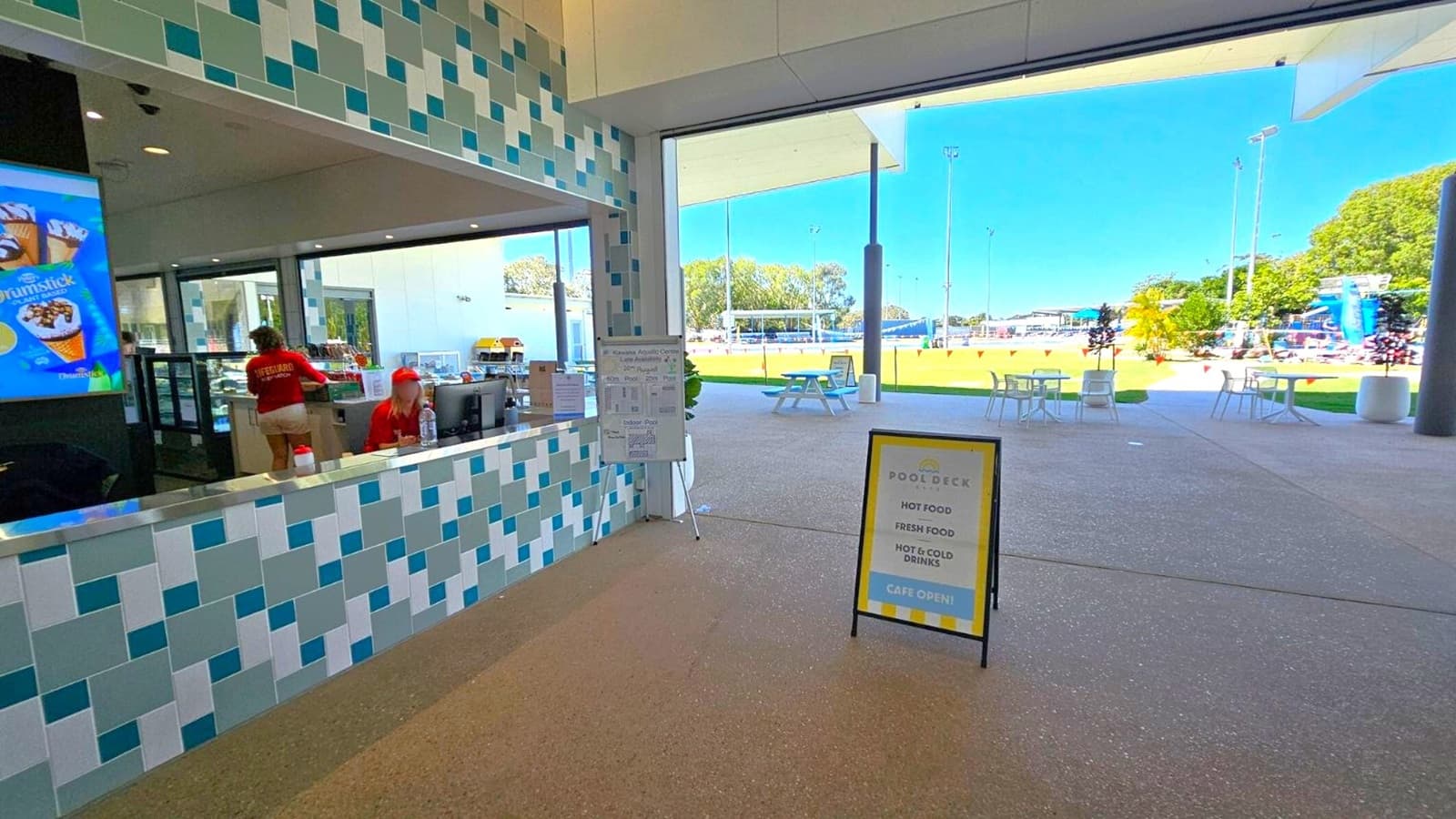 On the left, a counter with a blue and white tiled design, where a person in a red shirt is standing. Behind the counter, a digital display shows an ice cream advertisement. In the centre, a sign lists pool opening hours. On the right, a sandwich board advertises food and drinks. Beyond the open area, outdoor seating with tables and chairs is visible, along with a grassy field and sports equipment. The floor is a mix of brown and purple speckled surfaces.