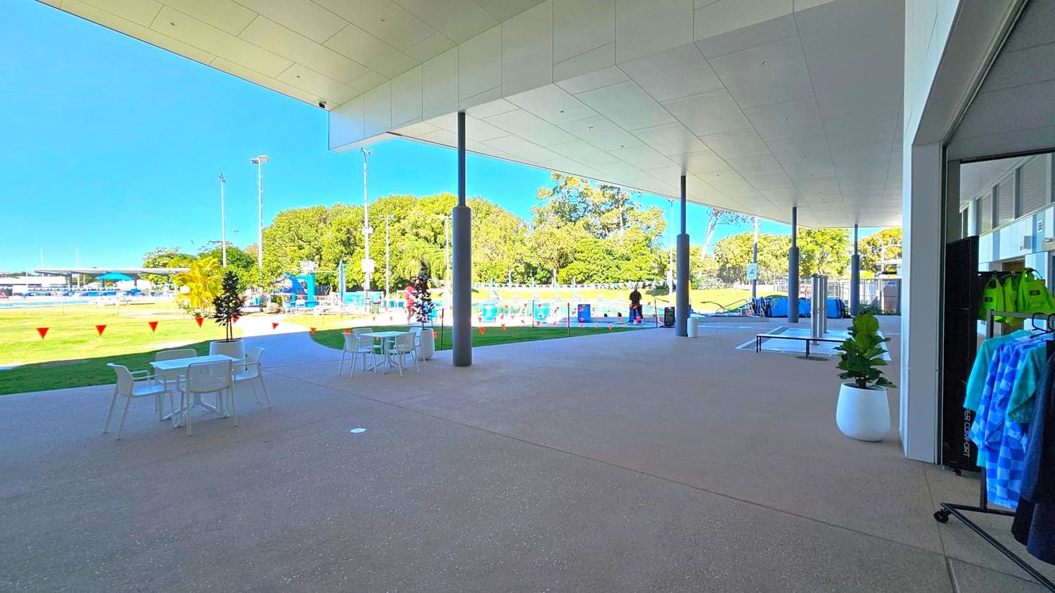 A covered outdoor area with a smooth, speckled floor and a ceiling with recessed lighting. On the right, a clothing rack with towels and a potted plant. In the middle, several white tables and chairs. Beyond the covered area, a grassy field with orange bunting, a swimming pool, and people in the distance. Tall trees and light poles are visible in the background.