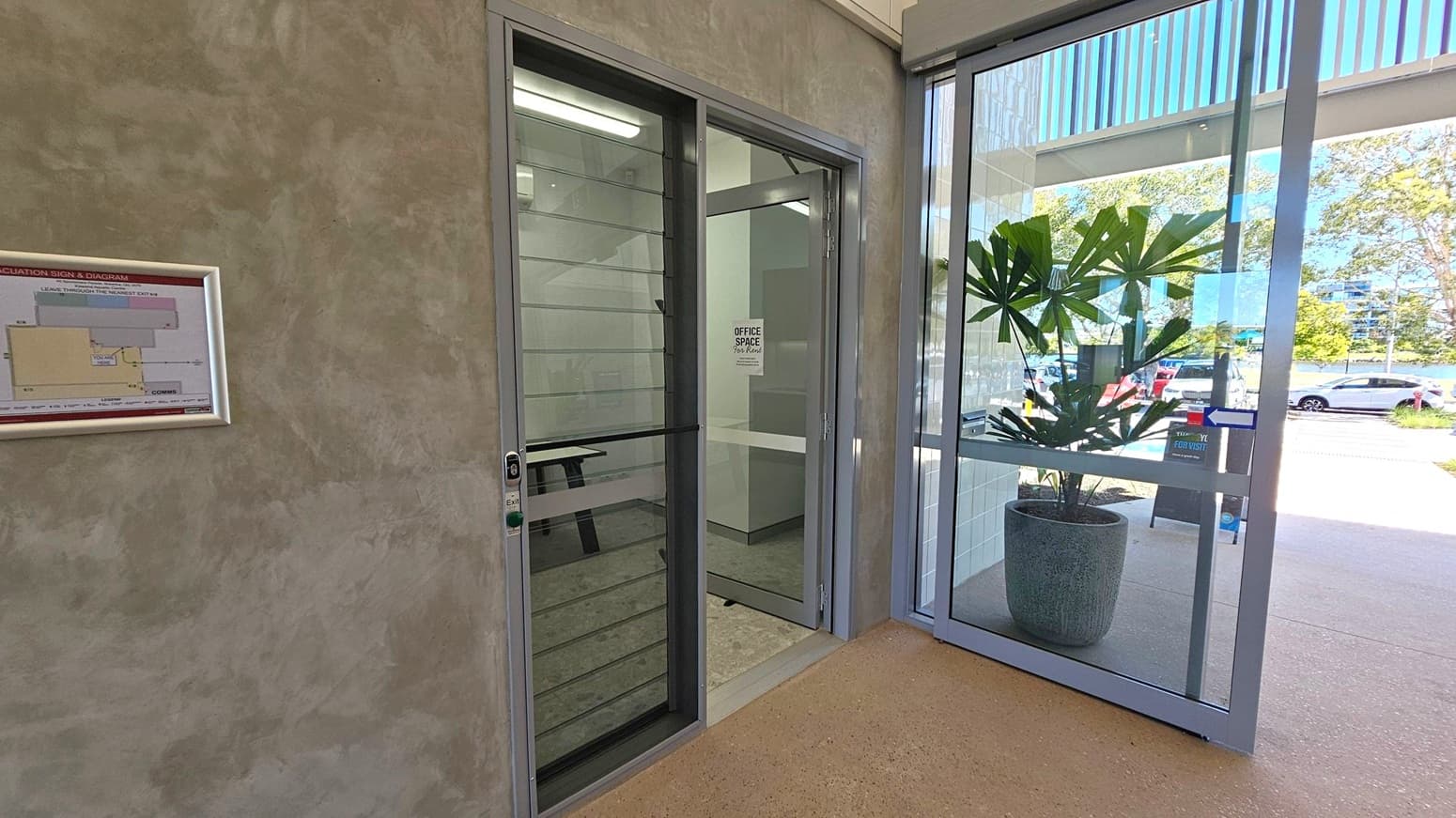 A corridor with a textured wall on the left featuring an evacuation sign and diagram. A glass door with a sign reading "Office Space" is next to it. On the right, a large glass window and door reveal a potted plant with broad leaves. Outside, there are parked cars and trees visible. The floor is a light brown, speckled surface.