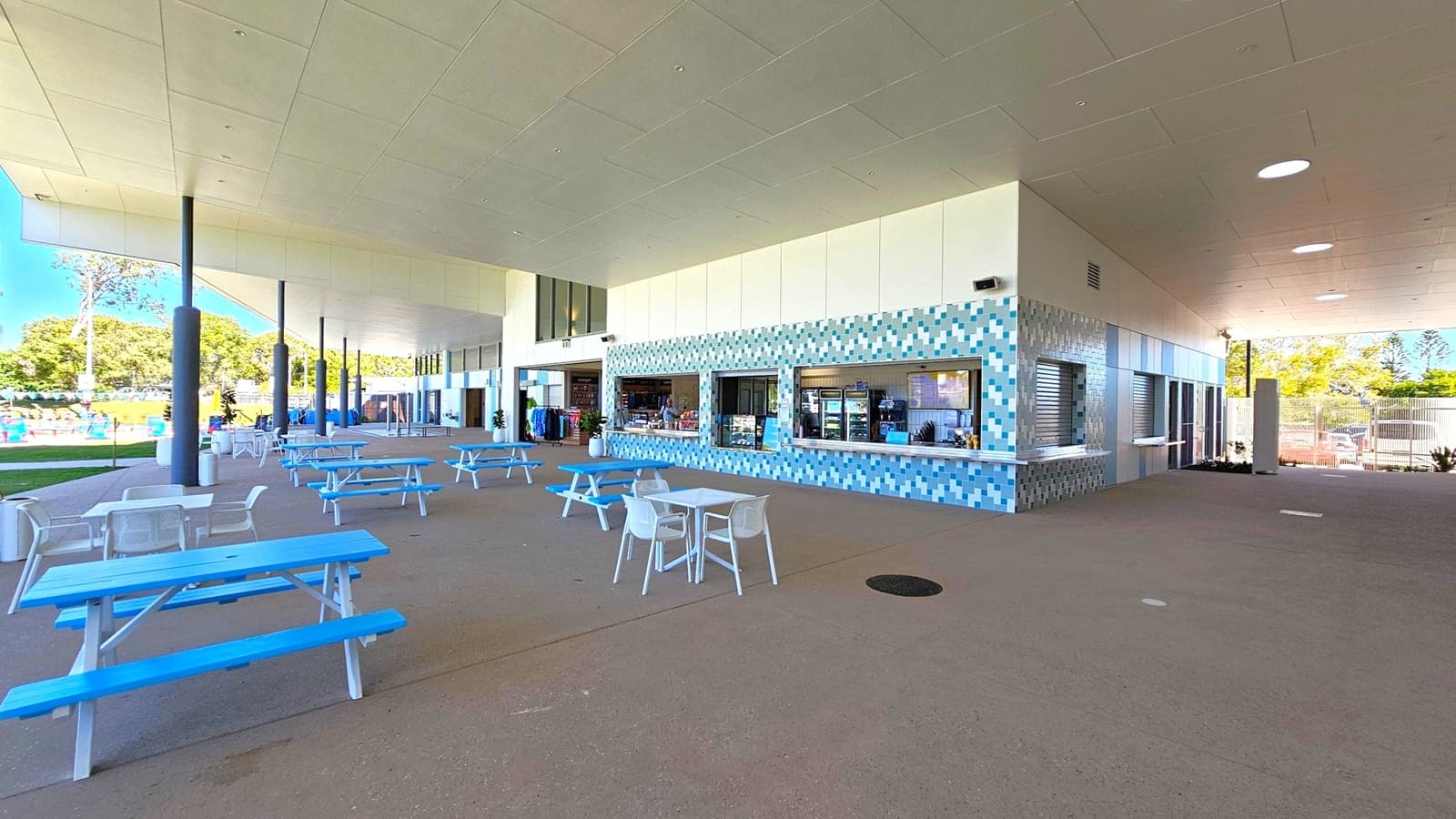 Outdoor seating area with blue picnic tables and white chairs on a concrete floor. On the right, a building with a façade of blue and white tiles features several service windows. The ceiling is white with circular lights, supported by black columns. In the background, a grassy area with trees and a glimpse of a swimming pool can be seen.