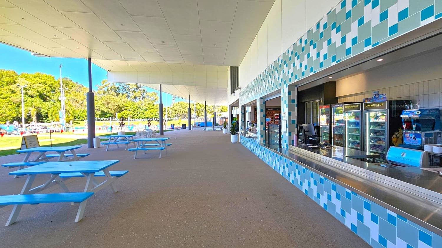 A covered outdoor area with a tiled wall on the right featuring a service counter and refrigerators displaying products. Blue and white picnic tables are arranged on the left. The ceiling is white and supported by columns. In the background, there is a grassy area with trees and a swimming pool, where people are visible. The floor is a smooth, grey surface.