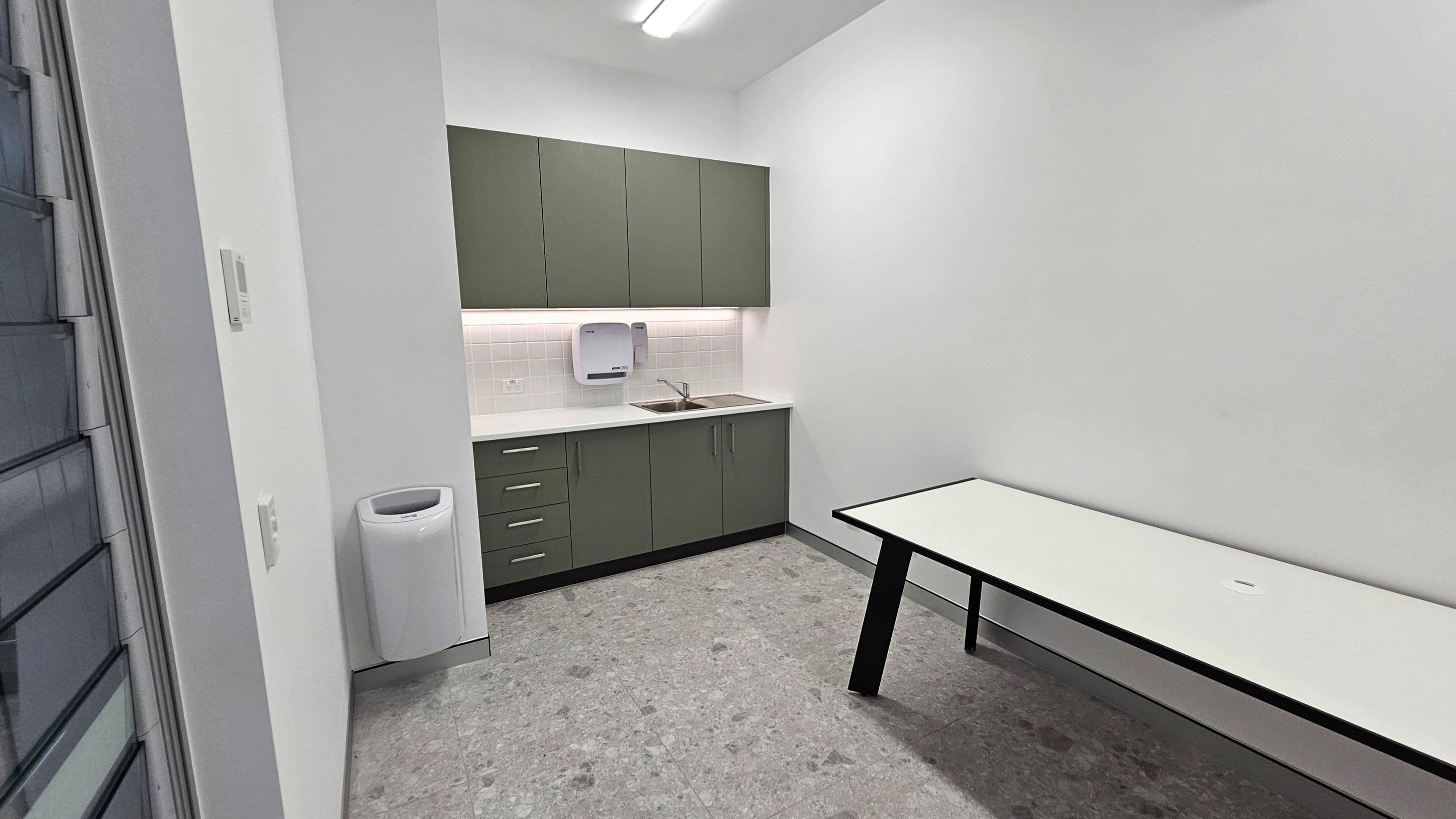 A small room with a kitchenette on the left, featuring green upper cabinets and grey lower cabinets. A white countertop holds a sink and a paper towel dispenser is mounted above. A white bin is positioned near the wall. On the right, a long white table with black legs extends along the wall. The floor is covered in grey tiles, and the ceiling has a fluorescent light fixture.