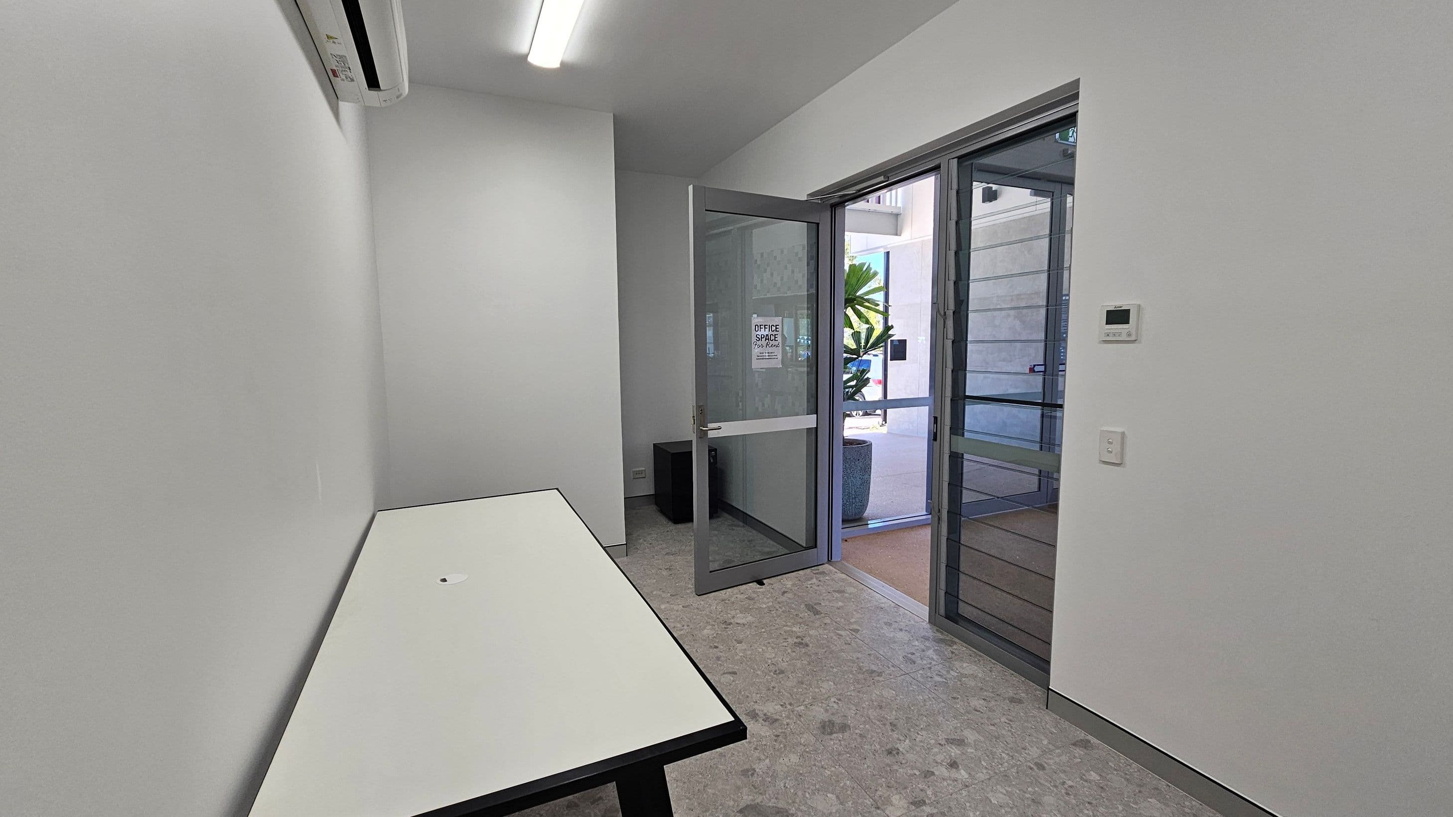 A small, narrow room with a white table on the left side. The floor is grey with a speckled pattern. On the right, a glass door is open, leading outside where a large potted plant is visible. Above the door, a fluorescent light fixture is mounted on the ceiling. An air conditioning unit is installed high on the left wall. A thermostat and a light switch are on the right wall. The walls are plain and white.