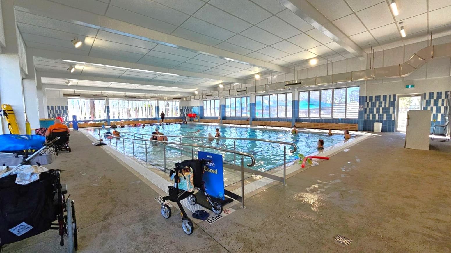 Indoor swimming pool with several people in the water. On the left, a person in a wheelchair and another wheelchair are visible. In the foreground, a walker is placed near a sign that reads "Lane Closed." The pool is surrounded by large windows on the right, allowing natural light to enter. The ceiling is lined with fluorescent lights, and the floor is a mix of wet and dry areas. Blue and white tiles decorate the walls.