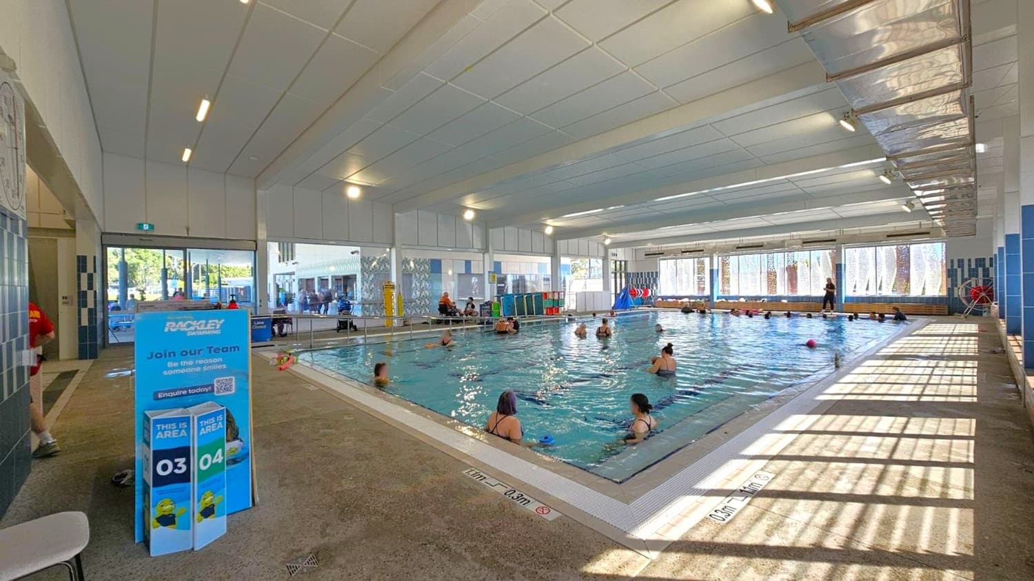 Indoor swimming pool with several people in the water, possibly taking a class. On the left, a blue sign with recruitment information stands near the entrance. The pool is surrounded by a tiled deck, and large windows on the right allow natural light to filter in, casting shadows on the floor. The ceiling is lined with panels and lights. In the background, there are more people and some pool equipment.