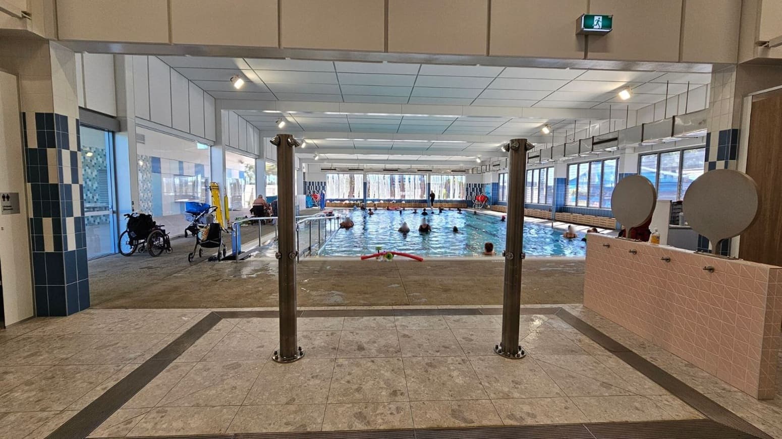 Indoor swimming pool with several people in the water. The pool is surrounded by large windows on the left and right, allowing natural light to enter. In the foreground, there are two wooden posts and a tiled floor with wet footprints. On the left, a wheelchair and a stroller are visible near the windows. On the right, there are two circular mirrors mounted on a tiled wall. The ceiling is white with recessed lighting, and an exit sign is visible above.