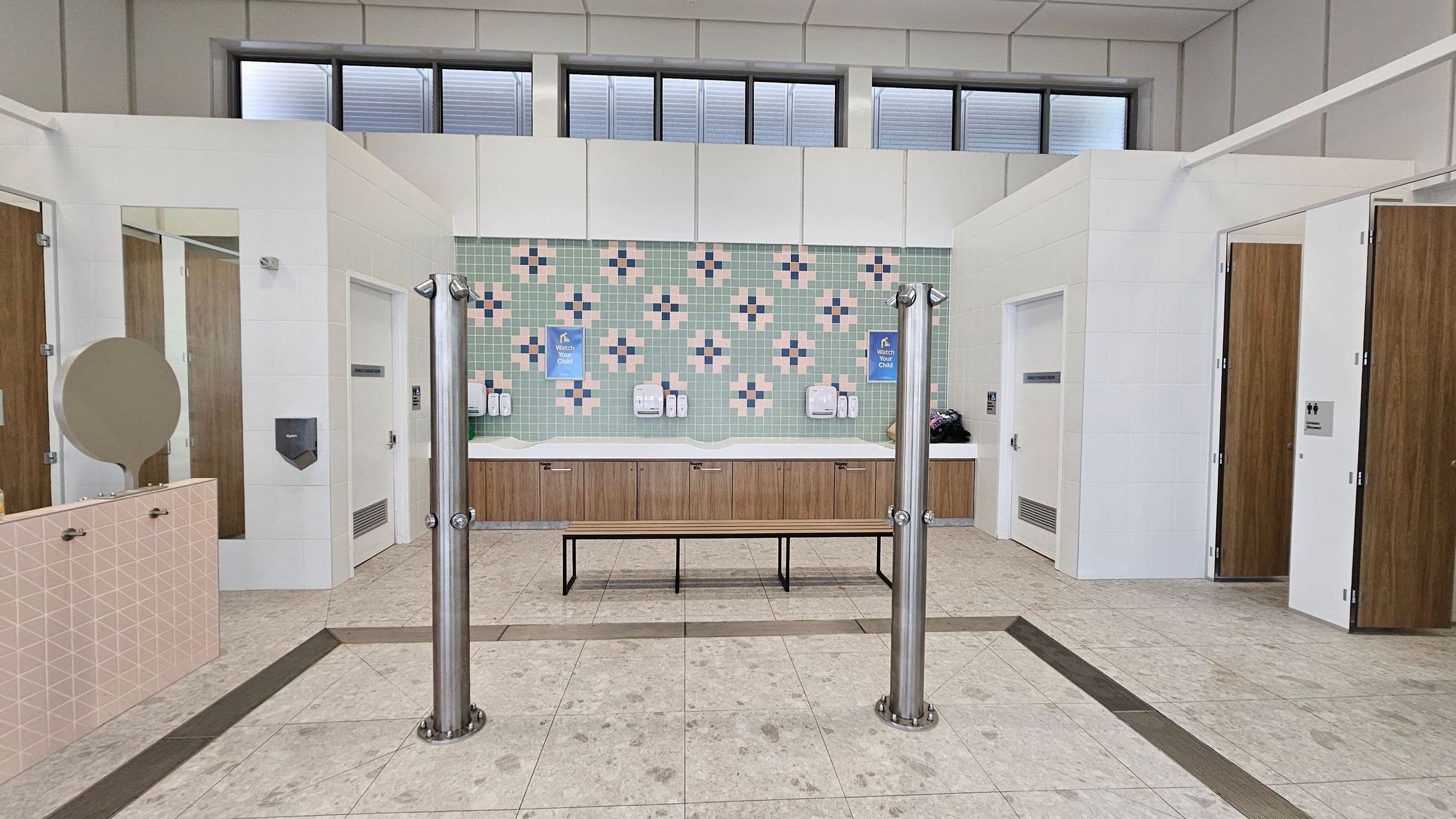 A public restroom with a tiled floor and high ceiling. In the centre, a bench is positioned in front of a wall with green and pink patterned tiles. Above the sinks, there are soap dispensers and signs. On the left, there are cubicles with wooden doors and a round mirror. On the right, more cubicles with similar doors. The upper part of the wall has windows with frosted glass panels. Stainless steel poles are positioned near the bench.