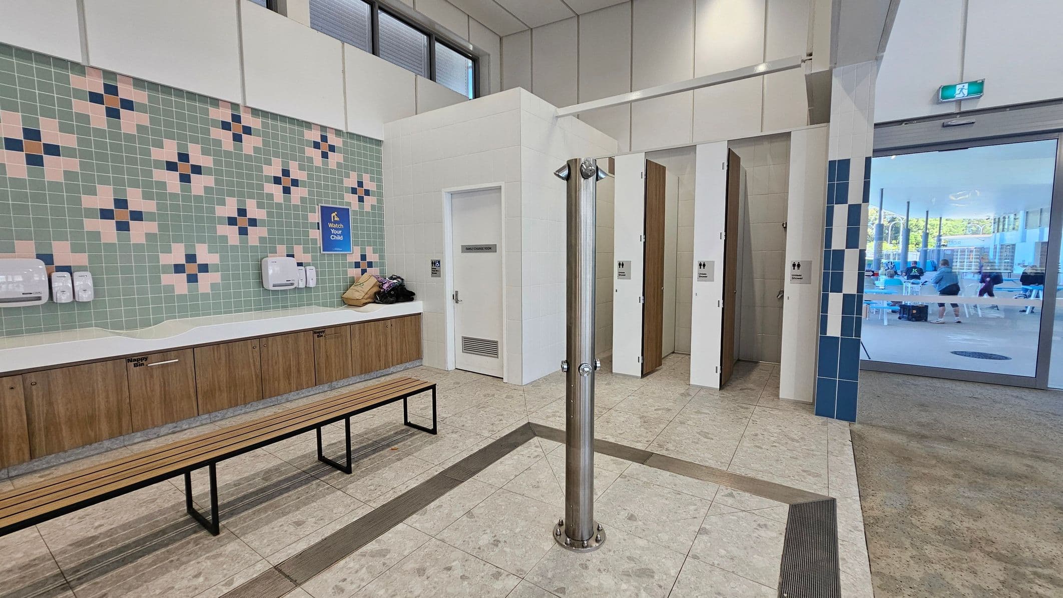 A changing room with a tiled floor and high ceiling. On the left, there is a wall with a mosaic tile pattern in blue and pink, featuring a long bench and a counter with dispensers and a nappy bin. In the centre, a metal shower pole stands on the floor. On the right, there are open cubicles with wooden doors. Beyond a glass door, people are visible in a seating area.
