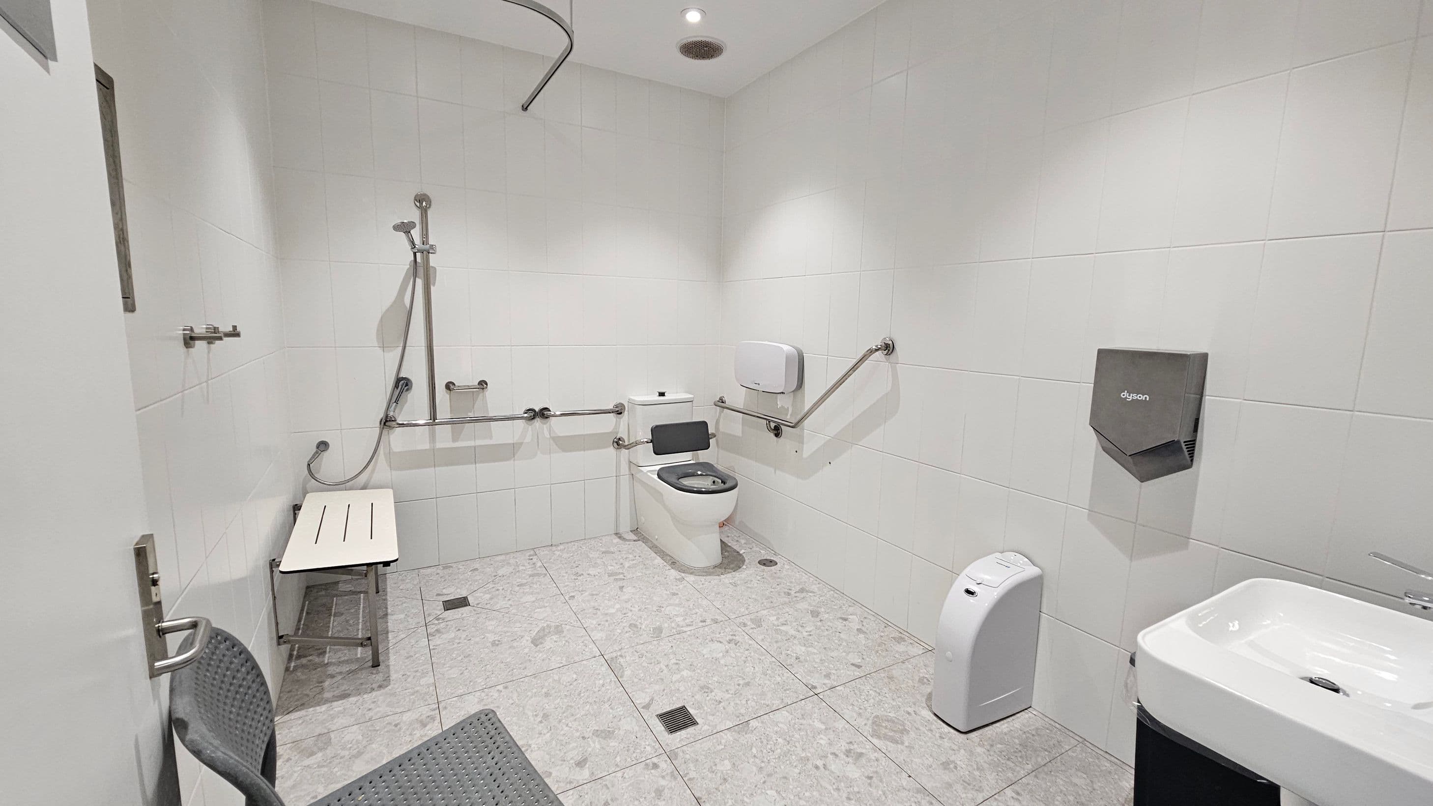 A spacious, accessible bathroom with white tiled walls and a light grey tiled floor. On the left, a shower area with a foldable bench and grab bars. In the centre, a toilet with additional grab bars and a paper dispenser. On the right, a hand dryer and a sink with a tap. The ceiling has recessed lighting.