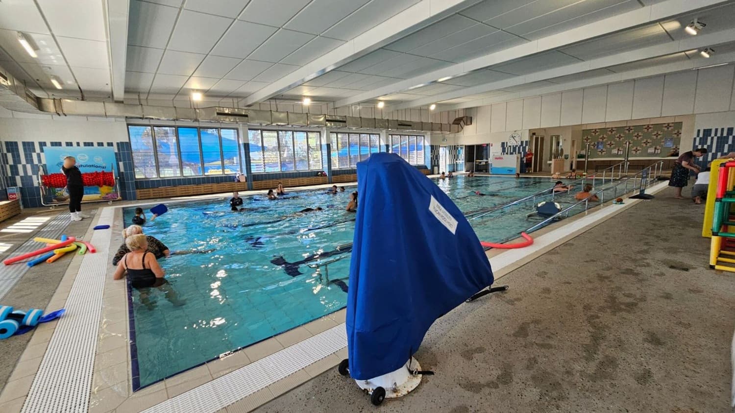 An indoor swimming pool with several people in the water, some using pool noodles. On the left, a person stands near a wall with a colourful display. A stack of pool noodles lies on the poolside. In the centre, a blue-covered object is positioned on the pool deck. The right side shows a ramp leading into the pool and a person bending over near the entrance. Large windows line the background, allowing natural light to illuminate the space. The ceiling is white with recessed lighting.