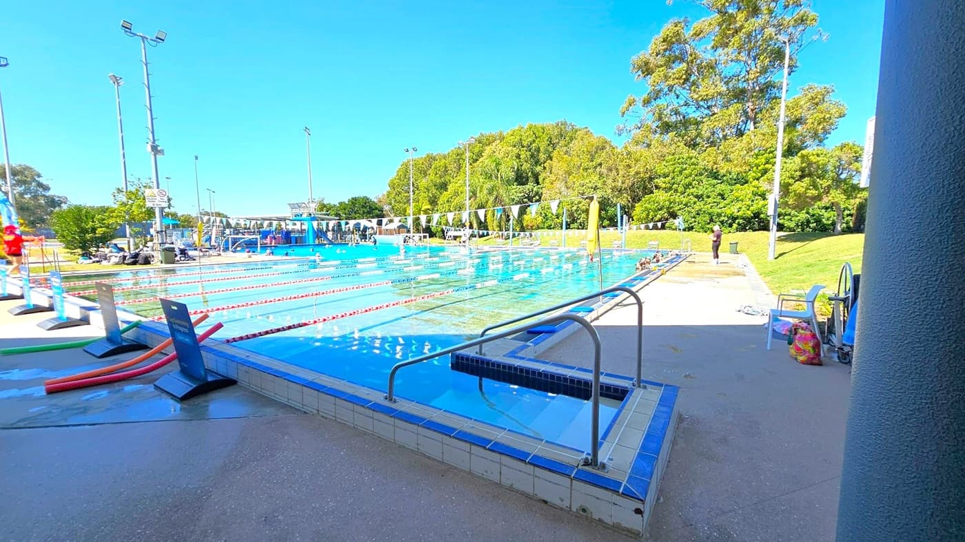 An outdoor swimming pool with multiple lanes, separated by red and white lane dividers. On the left, there are poolside signs and a few pool noodles. The right side shows a grassy area with trees and a person standing near the pool. In the foreground, a metal handrail leads into a smaller pool section. The background features tall trees and several floodlights. The pool deck is wet, with chairs and a colourful bag nearby.