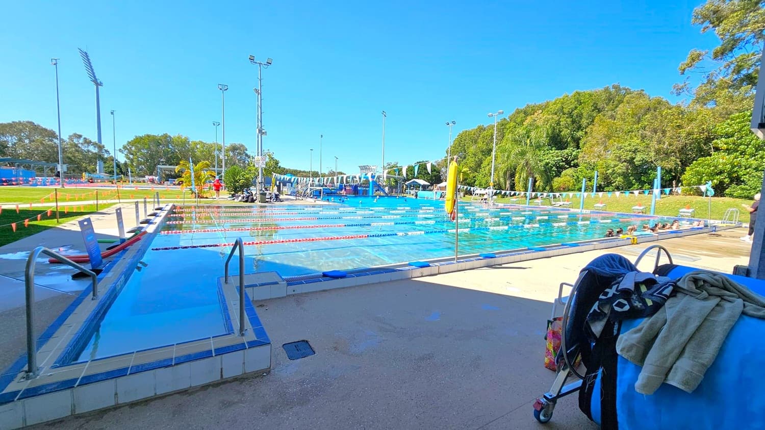 A swimming pool with multiple lanes is in the centre, surrounded by a paved area. On the left, there are metal handrails leading into a smaller pool section. In the foreground, a stroller with towels draped over it is visible. The background features lush green trees and several tall floodlights. Swimmers are in the pool, and a yellow lifebuoy is mounted on a pole near the poolside.