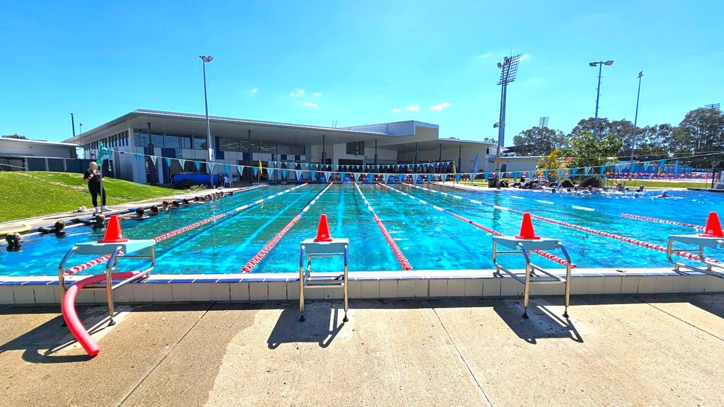 An outdoor swimming pool with clear blue water, divided into lanes by red and white lane markers. Orange cones are placed on the starting blocks at the pool's edge. On the left, a person stands near the poolside, while on the right, several people are in the water. In the background, a modern building with large windows and a flat roof is visible, along with tall floodlights and trees. The pool deck is concrete.