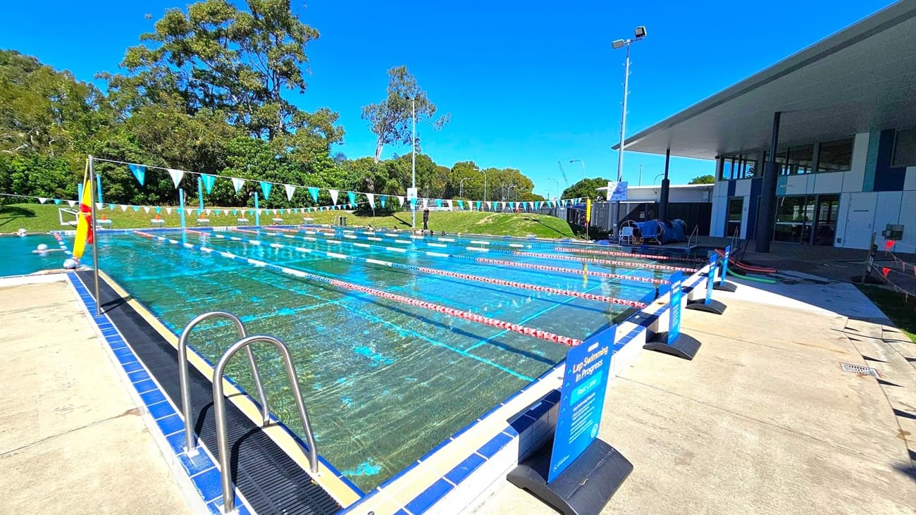 An outdoor swimming pool with multiple lanes, separated by red and white lane dividers. A metal ladder is on the left side, and a sign indicating "Lap Swimming in Progress" is on the right. Blue and white triangular flags are strung above the pool. In the background, there are trees and a grassy area. A building with a slanted roof is on the right, and the pool deck is made of concrete.