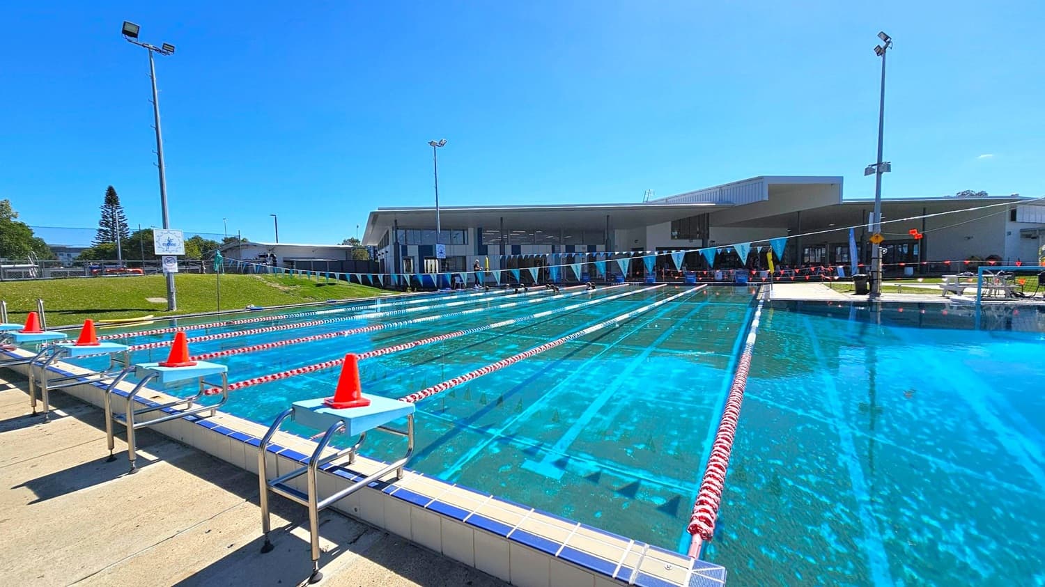 An outdoor swimming pool with multiple lanes, each separated by red and white lane dividers. Orange cones are placed on the starting blocks at the pool's edge. In the background, a modern building with large windows and a flat roof is visible. A grassy area with a tall tree and several light poles surrounds the pool. Blue triangular flags are strung across the pool. The pool deck is made of light-coloured tiles.