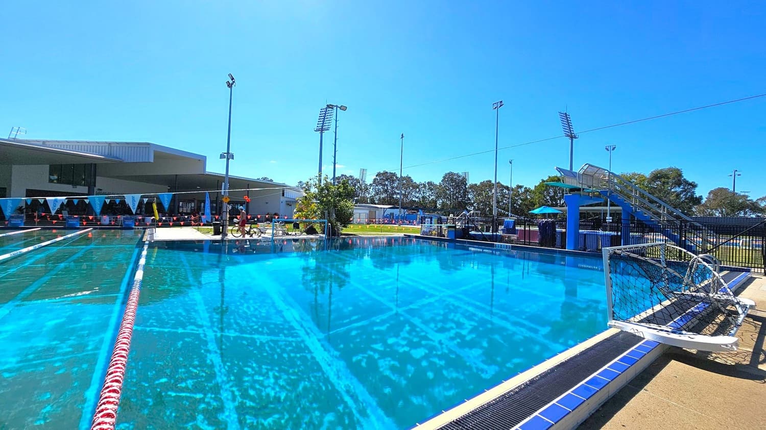 Outdoor swimming pool with clear blue water. On the left, there are lane dividers and a modern building with a shaded area. In the centre, a lifeguard sits on a chair near the pool's edge. On the right, a water polo goal is visible, along with a blue slide structure. Tall floodlights and trees are in the background. The pool deck is concrete, and the area is fenced.
