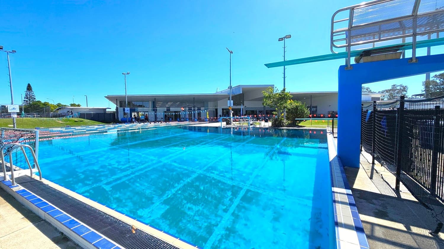 An outdoor swimming pool with clear blue water is in the foreground. On the right, a diving board is mounted on a blue structure. The pool is surrounded by a tiled deck and a black mesh fence. In the background, there is a modern building with large windows and a flat roof. Several tall floodlights are positioned around the pool area. A grassy area with a tree and a small building is visible on the left.