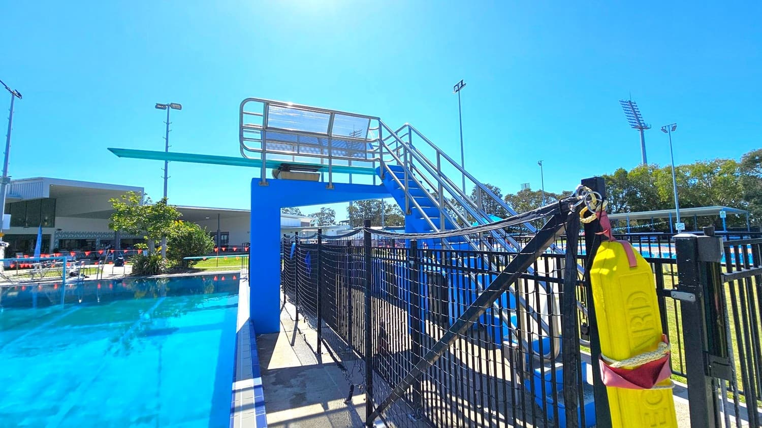 A diving board is positioned on the left, elevated on a blue platform with metal railings and stairs leading up to it. A swimming pool with clear blue water is in the foreground. On the right, a black fence with a yellow flotation device attached is visible. In the background, there are trees, a building, and tall floodlights. The area is well-lit, suggesting a sunny day.