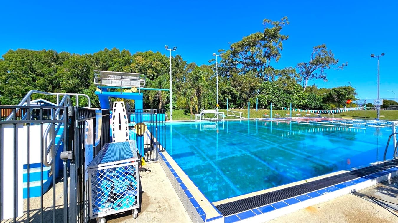On the left, there is a diving platform with metal railings and a blue structure. A cart with a mesh cover and various equipment is nearby. In the centre, a large swimming pool with clear blue water is visible, surrounded by tiled edges. On the right, there are trees and grassy areas with several tall floodlights. The background features more trees and a clear blue sky.