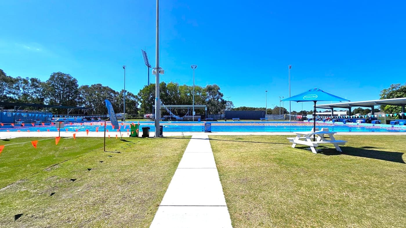 A swimming pool area with a concrete path leading to the pool in the centre. On the left, there are orange triangular flags and a blue flag on a pole. On the right, a white picnic table with a blue umbrella is on the grass. The pool is surrounded by blue and white bunting. In the background, there are trees and tall floodlights. The ground is a mix of grass and concrete.