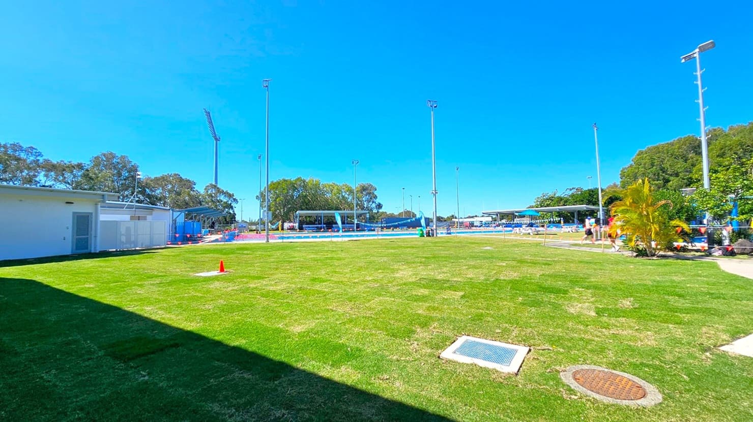 A grassy field with a white building on the left, partially in shadow. In the centre, several tall floodlights are visible, and a red cone is placed on the grass. To the right, there are small trees and a few people walking near a fenced area. In the background, more trees and structures are visible, including a covered seating area. The sky is clear and blue.