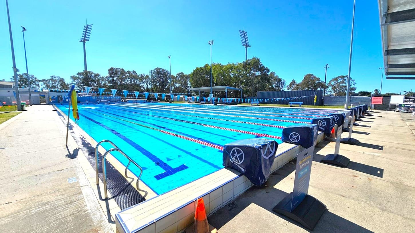 An outdoor swimming pool with multiple lanes, each marked by red and white lane dividers. Starting blocks are positioned at the pool's edge, covered with blue padding. A metal ladder is on the left side. The pool deck is concrete, with a traffic cone nearby. Tall floodlights and trees are visible in the background. A shaded seating area is on the far side, and a sign on the right reads "Lap Swimming in Progress."