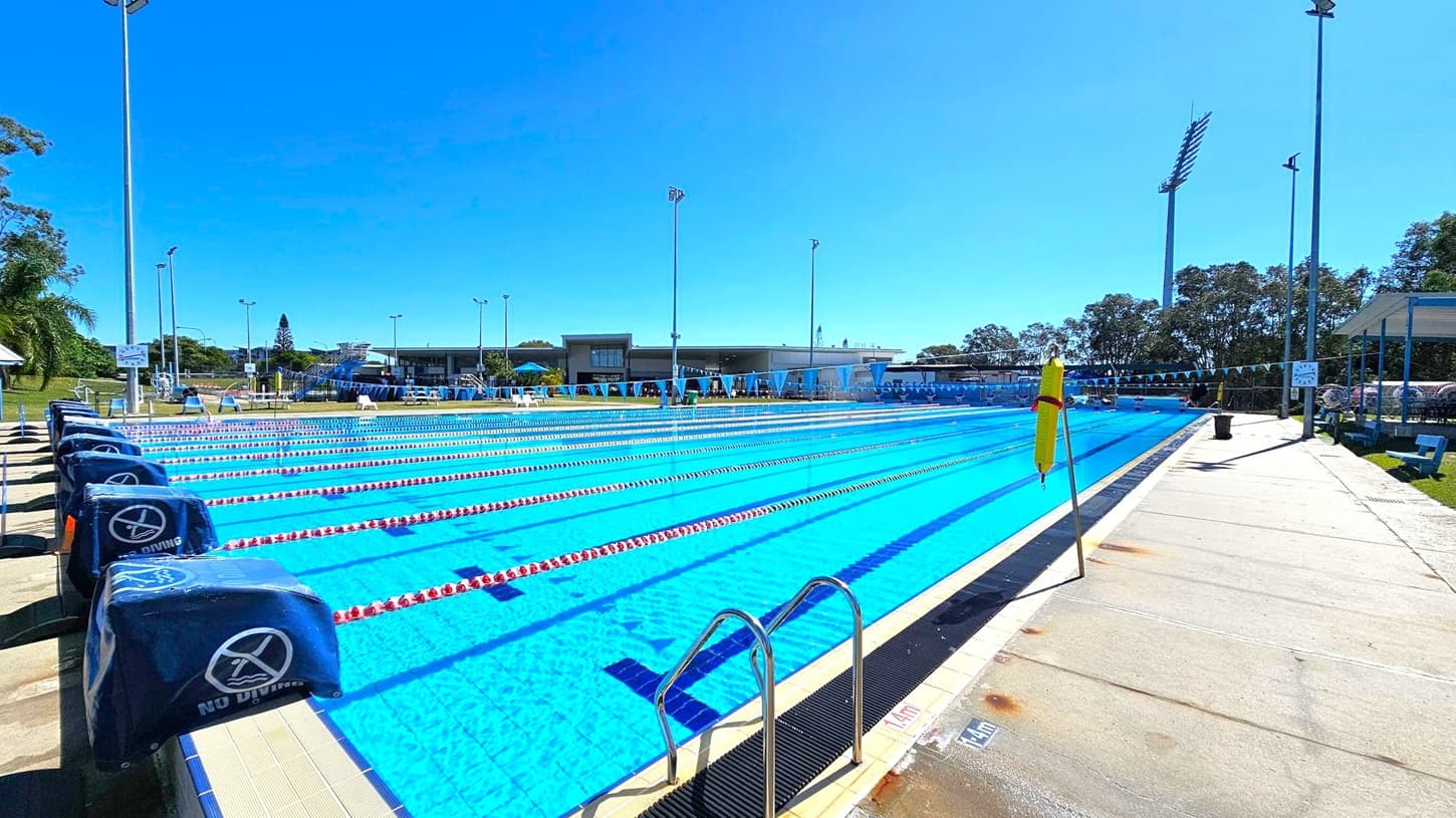 An outdoor swimming pool with multiple lanes, separated by red and white lane dividers. On the left, there are starting blocks covered with blue padding marked "No Diving." The poolside area is concrete, with a metal ladder on the right. In the background, there are trees, a building with a flat roof, and several tall floodlights. A yellow flag is positioned near the pool's edge. The area is well-lit, suggesting a sunny day.