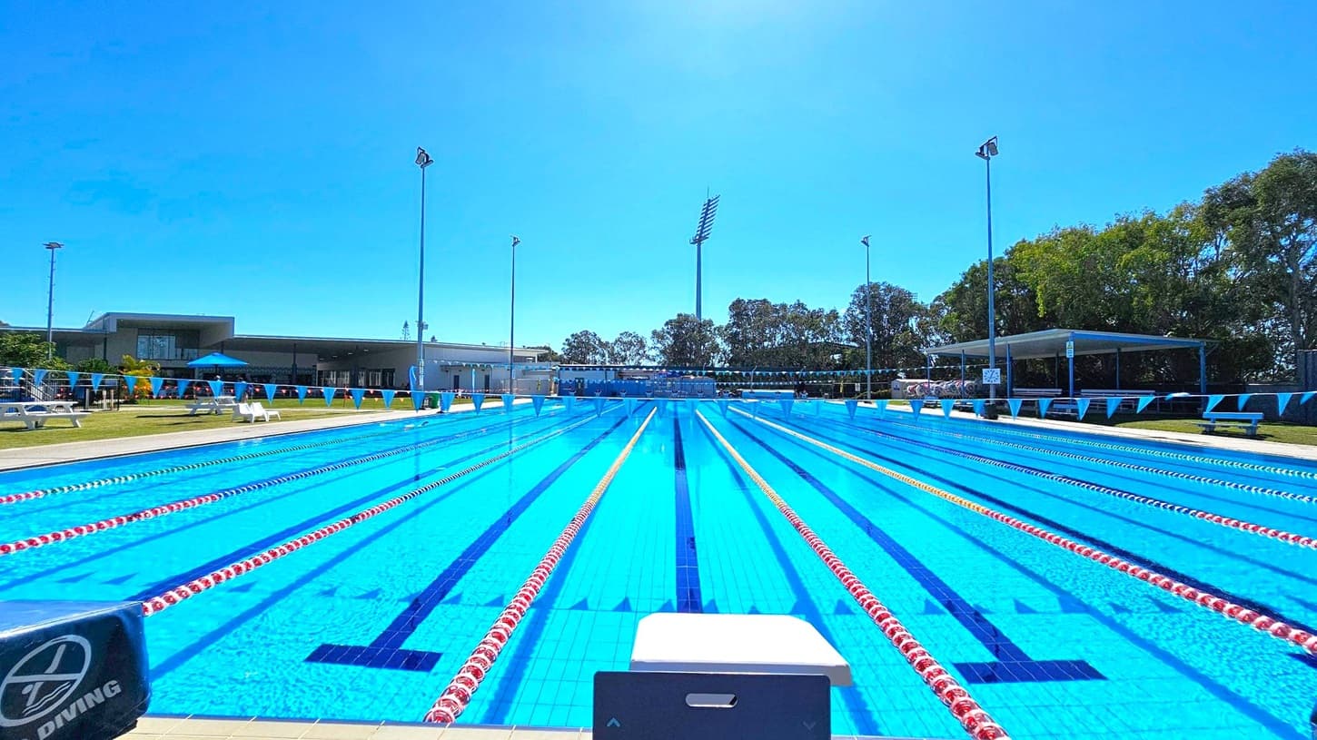 An outdoor swimming pool with multiple lanes, each marked by red and yellow lane dividers. A diving block is visible in the foreground. On the left, there are white benches and a building with a blue umbrella. On the right, trees and a shaded seating area are present. Tall floodlights are positioned around the pool, and a row of triangular flags stretches across the lanes. The pool area is surrounded by grass.