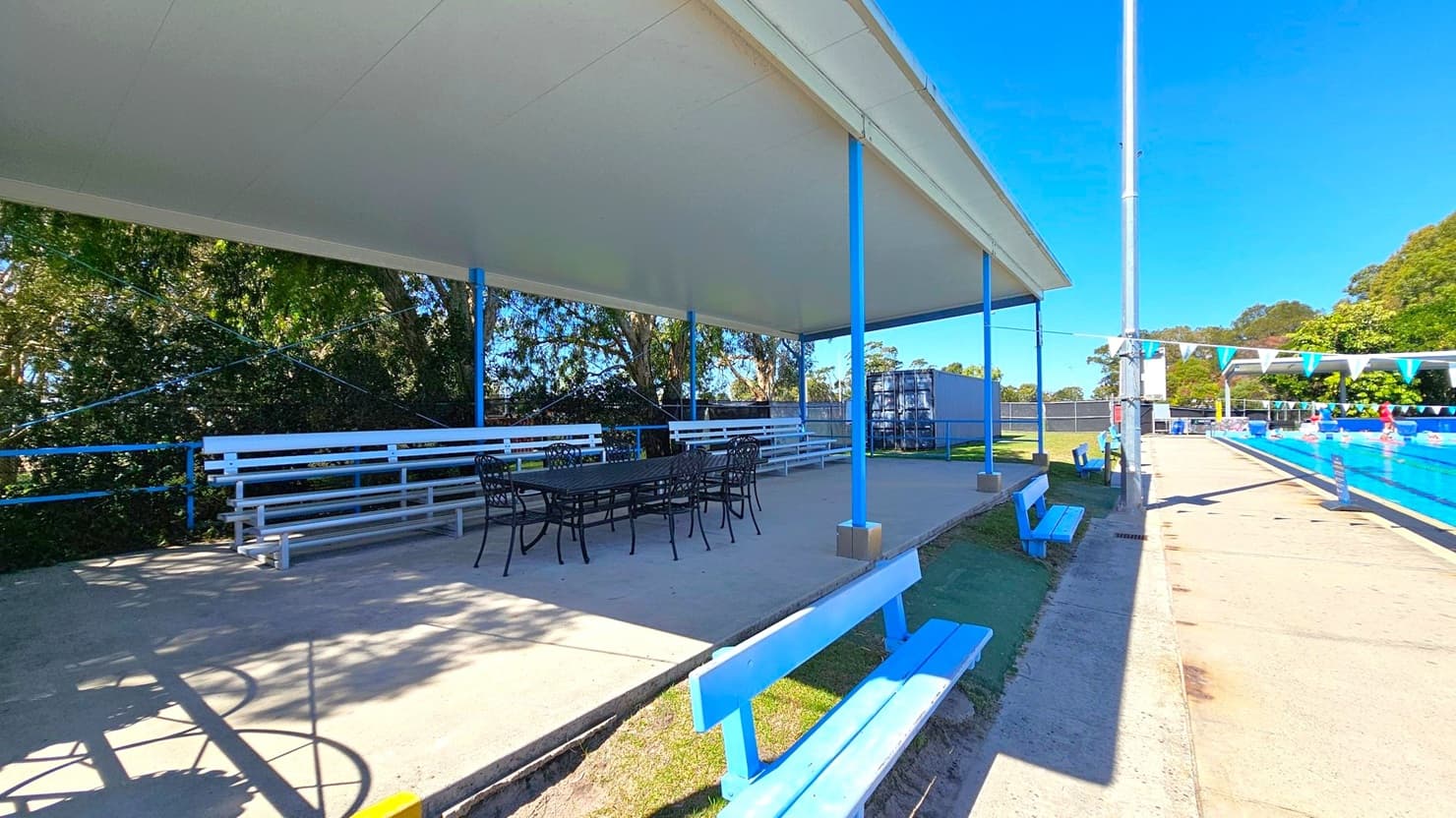 A covered seating area with white benches and a dark table with chairs is on the left. The area is supported by blue poles and surrounded by trees. On the right, a swimming pool with blue water is visible, with swimmers and lane dividers. Blue benches line the poolside, and a tall pole stands nearby. The ground is a mix of concrete and grass, and the ceiling of the covered area is plain.