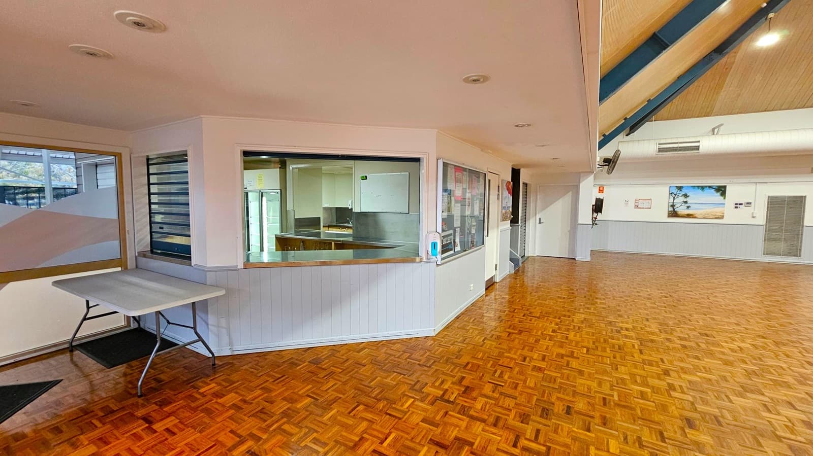A spacious hall with a high, triangular wooden ceiling and parquet flooring. At the far end, there's a stage with a wooden front and dark curtains. The walls are lined with large windows featuring frosted glass with geometric patterns. Ceiling lights and air ducts are visible, and there are speakers and a projector mounted above. The room is empty, providing an open, unobstructed view of the entire space.