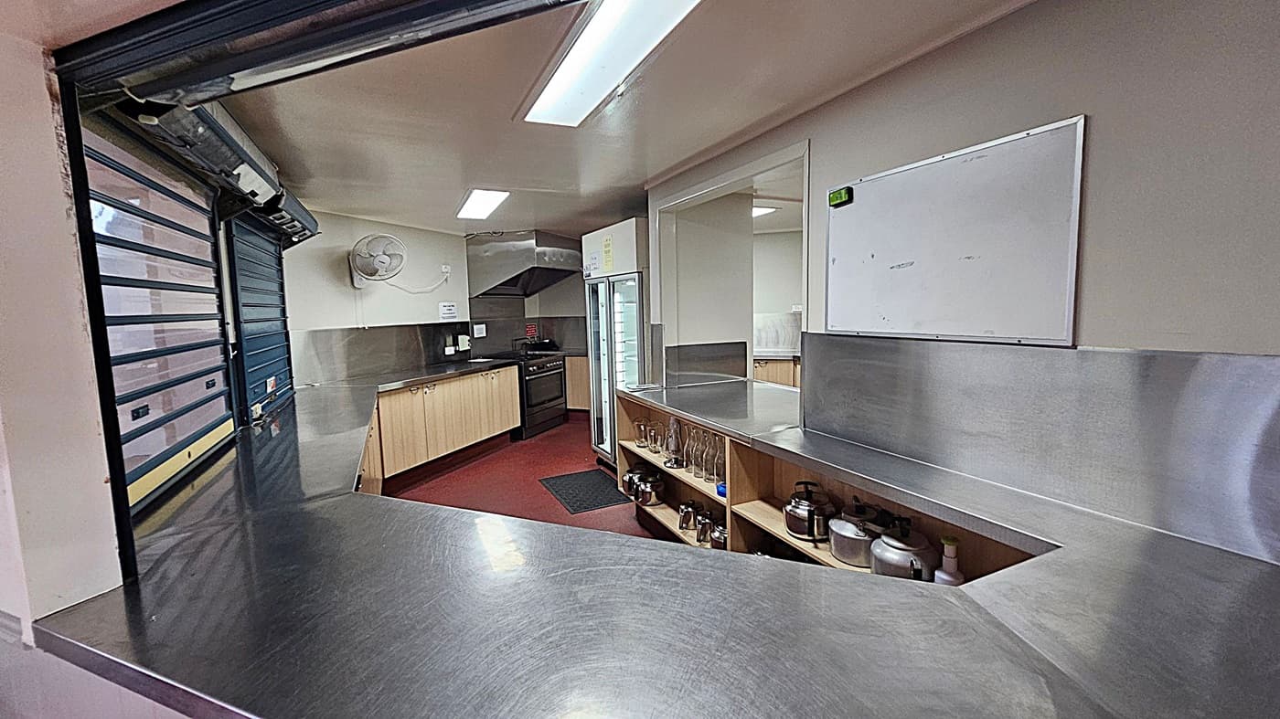 A commercial kitchen with stainless steel counters and a serving hatch. On the left, there's a closed roller shutter. In the middle, a fan is mounted on the wall above wooden cabinets and a black oven. A whiteboard is on the right wall. Below it, shelves hold glassware and kitchen utensils. A refrigerator with a glass door is next to the shelves. The floor is red, and the ceiling has fluorescent lights.