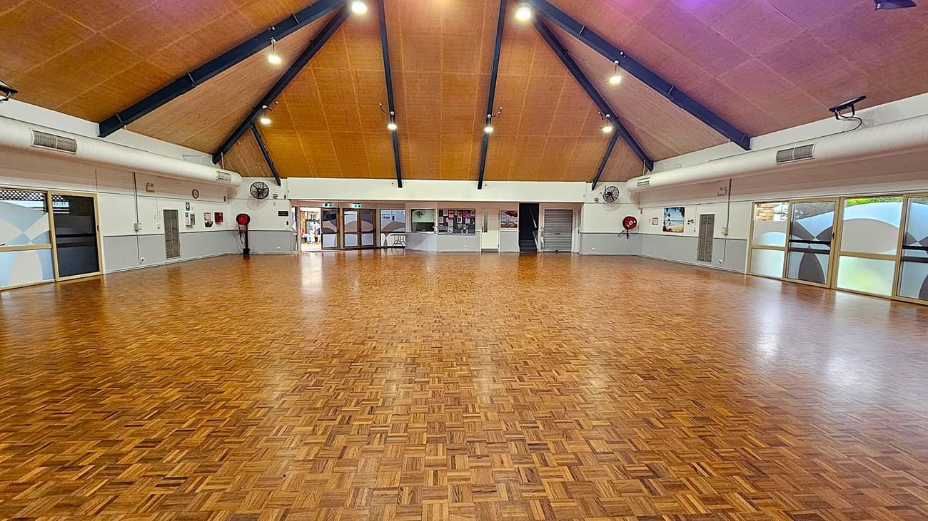 A spacious hall with a polished parquet wooden floor. The ceiling is high and triangular with exposed beams and multiple lights. The walls are light-coloured with several doors and windows, some with frosted glass. There are wall-mounted fans and fire safety equipment visible. A table is placed against the far wall, and there are various notices and posters displayed. The room is well-lit and appears to be a multi-purpose space.