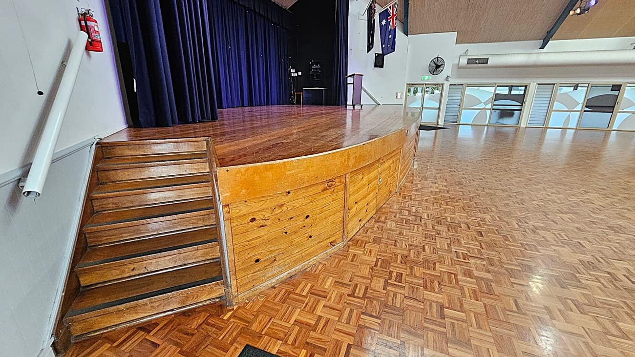 A wooden stage with a curved front is on the left, accessible by a set of wooden steps. A dark blue curtain hangs at the back of the stage. A fire extinguisher is mounted on the wall near the steps. The floor is a polished parquet pattern. On the right, there are large windows with frosted glass panels and a wall-mounted fan. The ceiling is high with visible beams. Flags hang on the wall above the stage.