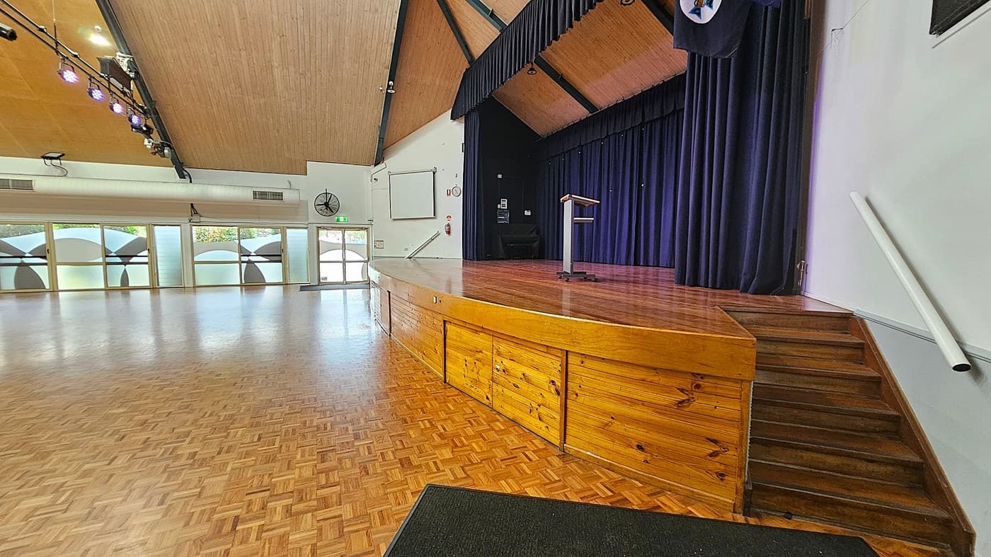 A spacious hall with a wooden parquet floor and a high, angled wooden ceiling. On the right, a stage with a wooden front and steps leading up to it, featuring a podium and dark curtains. On the left, large frosted windows with circular patterns allow natural light in. Ceiling lights are visible, and there's a wall-mounted fan and an exit door at the back.