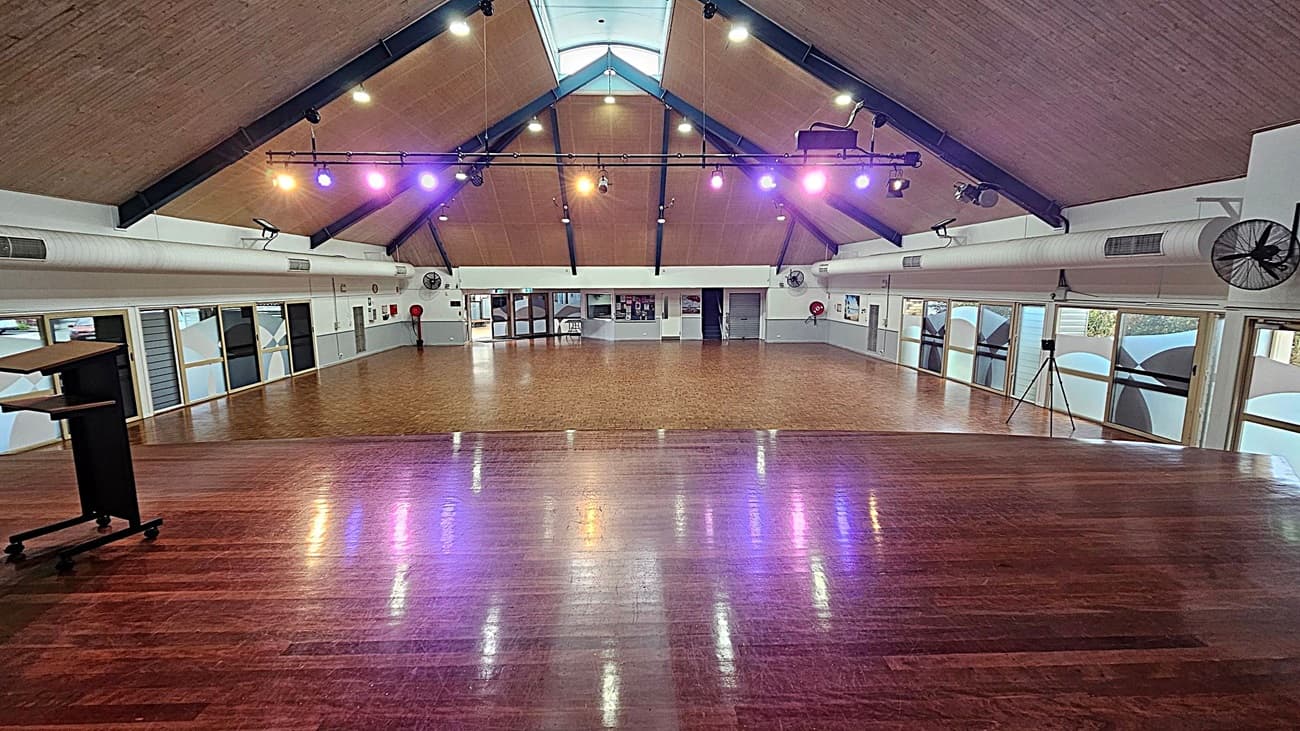A spacious hall with a high, triangular wooden ceiling and exposed beams. The floor is polished wood, reflecting coloured lights from above. On the left, there's a podium on wheels. The walls have large windows with frosted designs, and there are air conditioning ducts and fans mounted high. At the far end, there are doors and a small stage area. The lighting is a mix of warm and cool tones, creating a vibrant atmosphere.