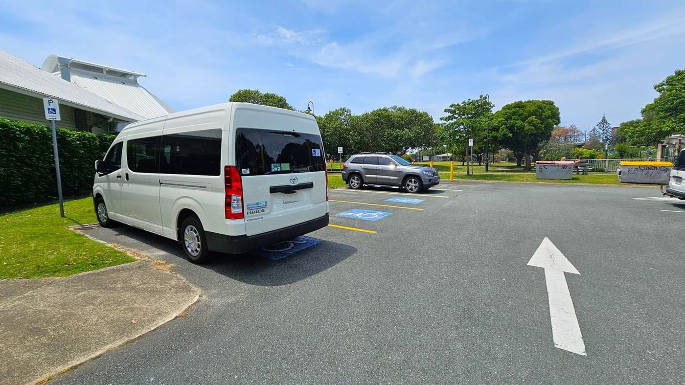A white van is parked in a disabled parking space on the left, next to a building with a metal roof. A grey SUV is parked in another disabled space to the right. The ground is asphalt with marked parking spaces and a white directional arrow pointing forward. In the background, there are trees, a grassy area, and concrete barriers with graffiti.
