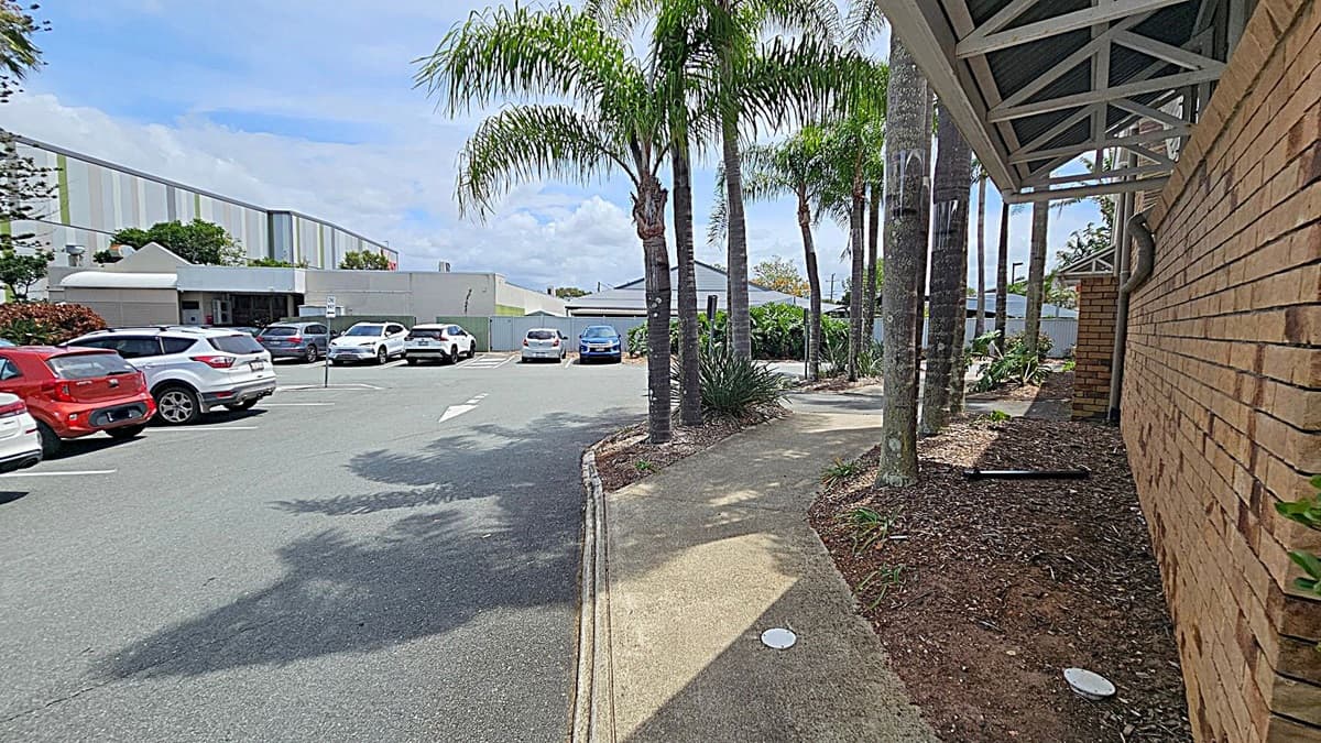 A car park with several vehicles, including a red and a white car, is on the left. In the centre, a row of palm trees lines a pathway. On the right, a brick building with a metal awning is visible. The ground is a mix of concrete and mulch with some plants. The background features a large building with a striped facade and more greenery.