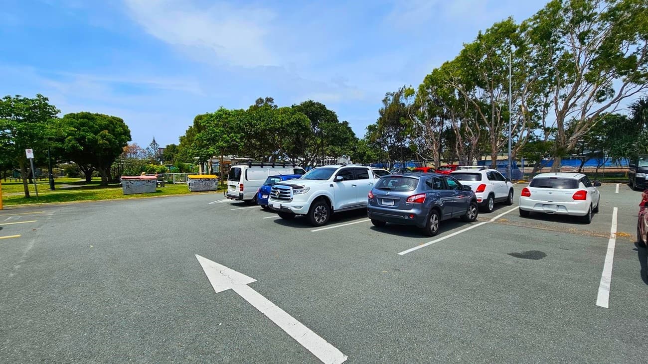 A car park with several vehicles, including a white SUV, a blue car, and a white van. The ground is asphalt with white parking lines and a directional arrow. In the background, there are green trees and a grassy area with a few large bins. The sky is clear, and the scene is well-lit, suggesting daytime.